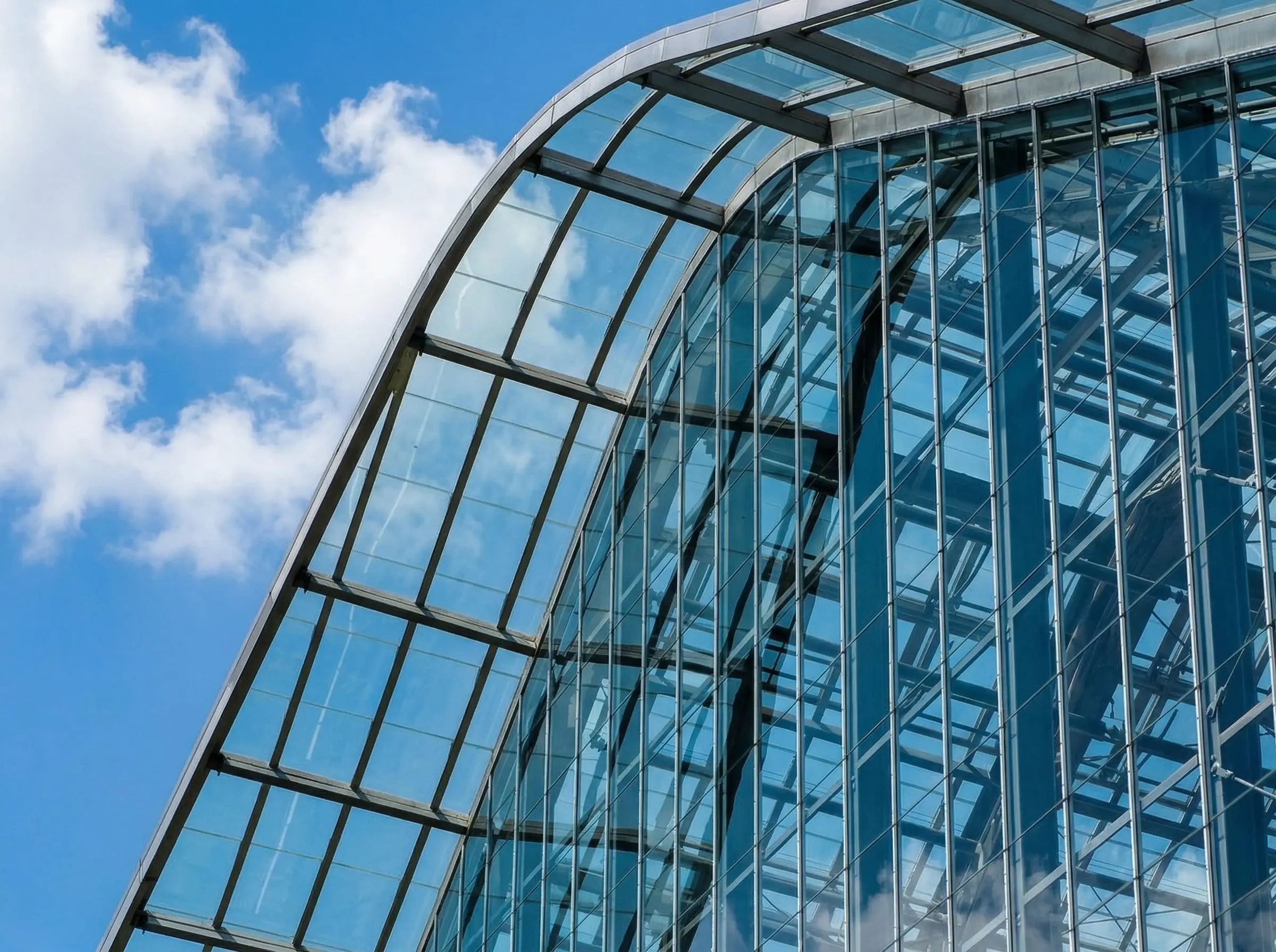 Фрагмент стеклянного фасада современного здания с металлическим каркасом и прозрачной кровлей на фоне голубого неба