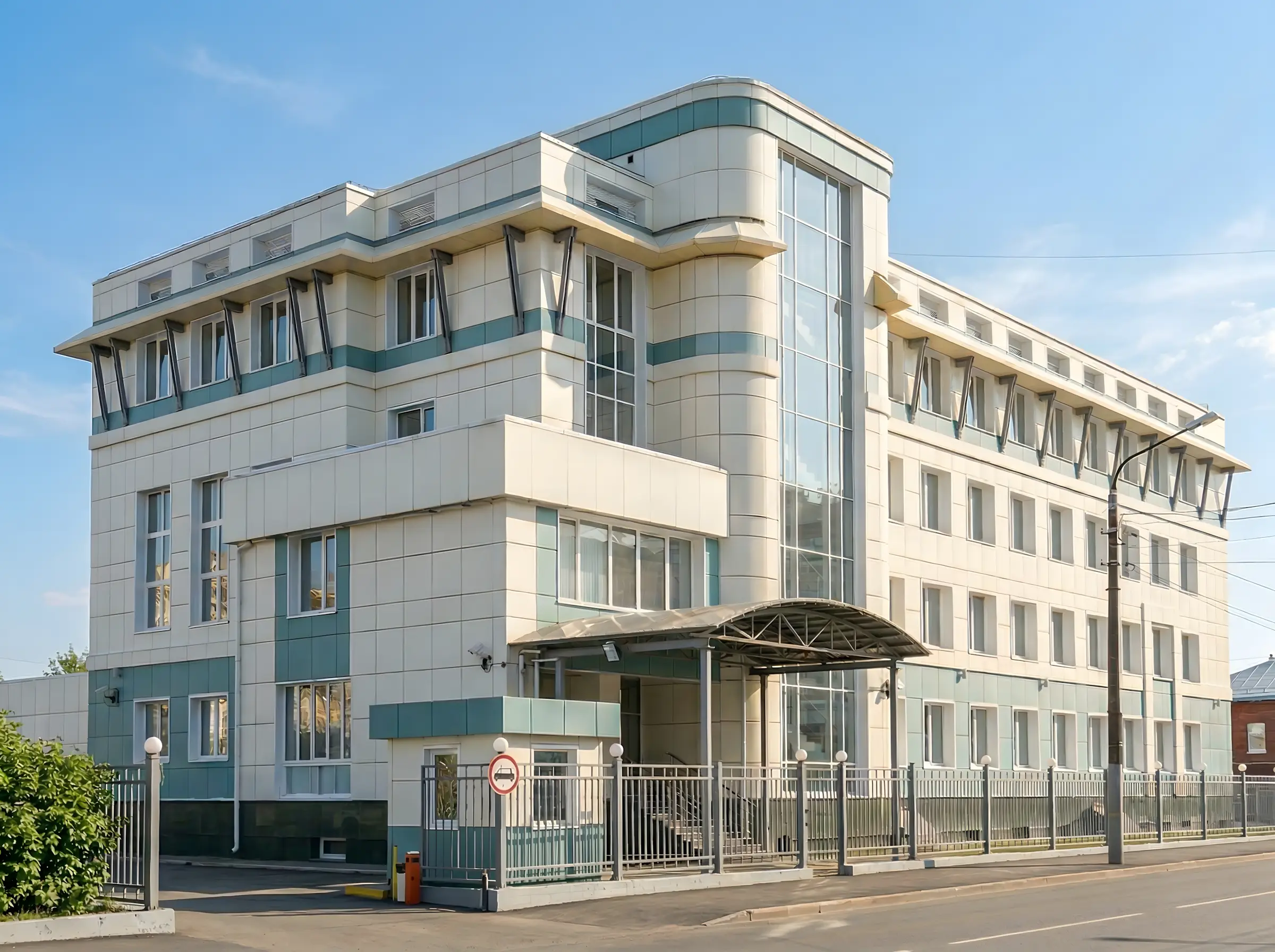 Четырехэтажное современное здание с остеклённым фасадом и металлическим забором, вход с козырьком, солнечная погода